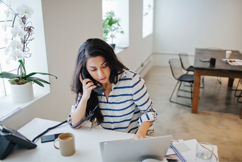 Photo of a woman in an office talking on a corded landline while entering notes into her laptop