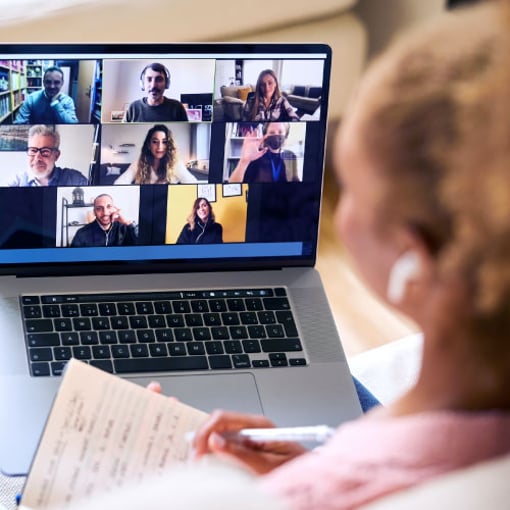 Video API infographic thumbnail Image of a video conference meeting on a laptop with multiple participants in small video squares; in front of the laptop and blurred out is a woman with a notebook in her left hand and a pen in her right hand to take notes during the meeting