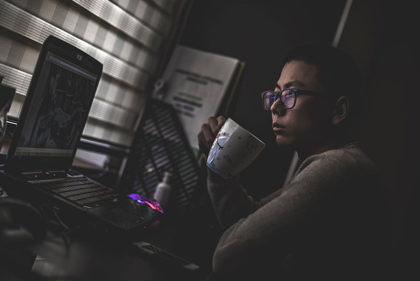 Side view of Asian man working remote from his laptop in a darkly lit room