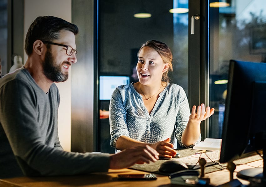 man and woman sitting at a desk and talking in front of a computer screen