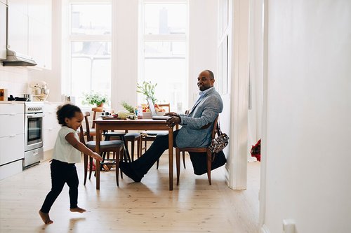 man in suit doing remote work with laptop from kitchen table while child runs by